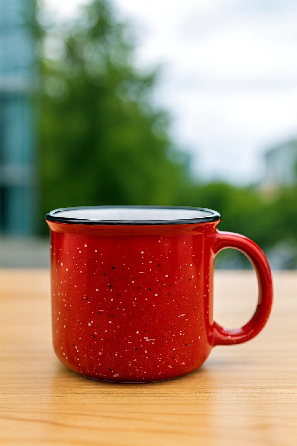 Red ceramic campfire mug with white speckles on a wooden table, set against a blurred outdoor background in Vancouver, ideal for branded swag giveaways.