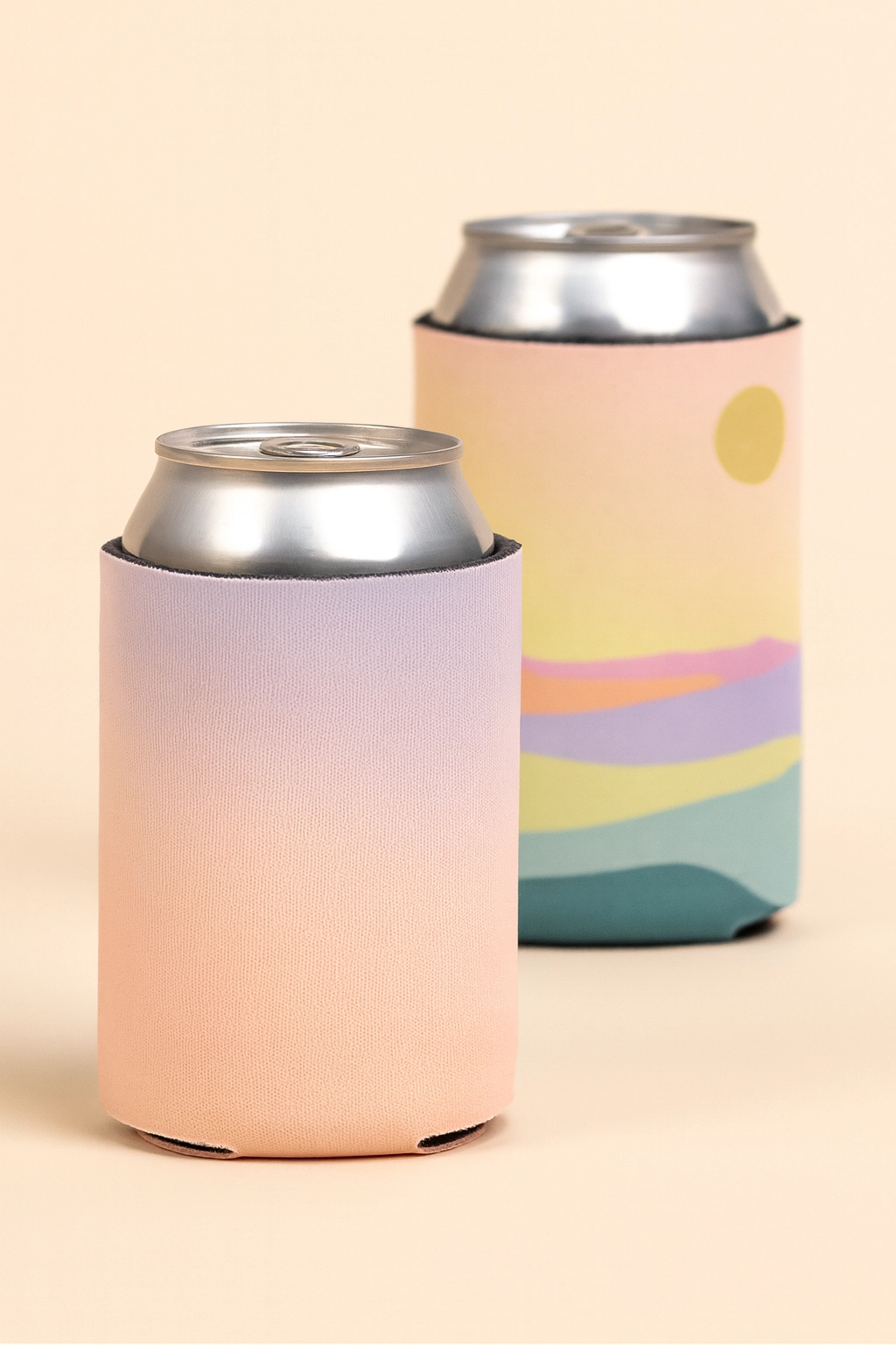 Two aluminum cans wrapped in pastel-colored koozies, one with a soft gradient design and the other featuring abstract landscape artwork, placed on a neutral beige background